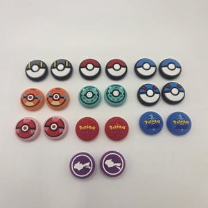 May include: A collection of colorful controller thumb grips featuring various designs. The grips include red, blue, orange, and green colors, with designs like Poke Balls and text that reads "Pokemon Shield".