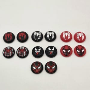 May include: A collection of twelve circular silicone thumbstick covers for game controllers, featuring various graphic designs including red and white spider emblems, black and white spider motifs, and character faces with sharp teeth and tongues.