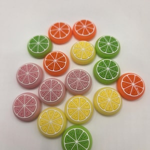 May include: A collection of colorful, circular fruit-shaped embellishments. The assortment includes orange, green, yellow, and pink slices, each with a white interior and a textured rind. These decorative pieces are arranged on a white surface.