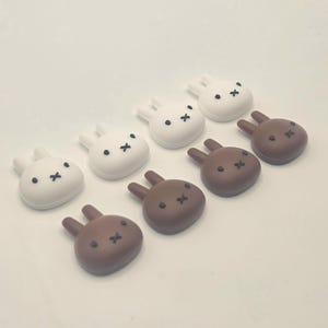 May include: An assortment of white and brown bunny-shaped objects. The white bunnies have black dot eyes and a black cross for a mouth. The brown bunnies have black cross eyes and mouths. The objects are arranged in rows.