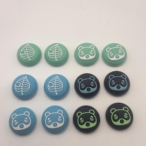 May include: A collection of twelve round controller thumb grips in various colors. The grips feature leaf and panda designs in white, light blue, and green. The grips are designed for video game controllers.