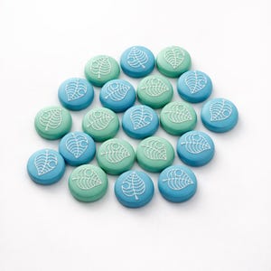 May include: A collection of round, teal and blue buttons with a white leaf design. The buttons are arranged on a white surface. The leaf design is a stylized outline of a leaf.