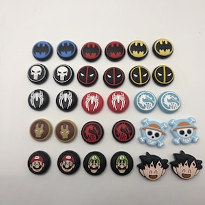 May include: A collection of black, red, blue, and yellow controller thumb grips featuring various comic book and video game characters. Designs include Batman, Spider-Man, Deadpool, and Mario.