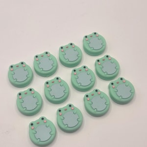 May include: A collection of twelve frog-shaped embellishments. Each frog has a light green body, a light blue belly, and pink cheeks. The frogs are arranged on a white surface.