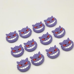 May include: A collection of eleven purple Gengar character charms, each featuring a wide white grin, pointed ears, and narrow red eyes. These small, flat, rubberized accessories are arranged in a loose grid pattern on a plain white surface.