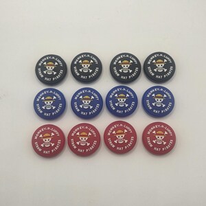 May include: A collection of twelve round buttons featuring the text "MONKEY.D.LUFFY STRAW HAT PIRATES" and a skull and crossbones design. The buttons are in black, blue, and red.