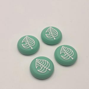 May include: Four teal-colored, round buttons with a white leaf design. The buttons are arranged on a white surface. The leaf design is a stylized, open-veined leaf.