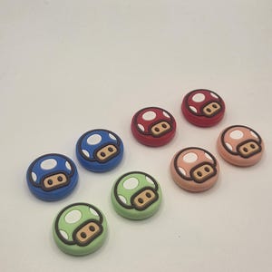 May include: A collection of colorful mushroom-shaped accessories. The accessories feature a white-dotted cap design in various colors, including blue, green, red, and pink. Each mushroom has a brown face.