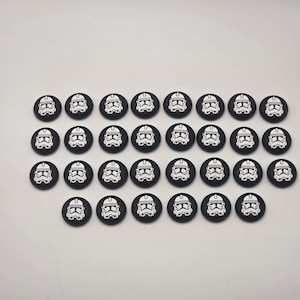 May include: A collection of thirty circular black buttons, each featuring a white graphic print of a stylized helmeted soldier character. The buttons are arranged in four rows, with the top three rows containing eight buttons each and the bottom row containing six.