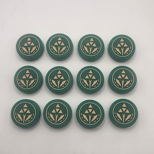 May include: Twelve green round objects with a beige geometric design. The objects are arranged in three rows of four. The design features a stylized triangle and lines. The objects are on a white surface.