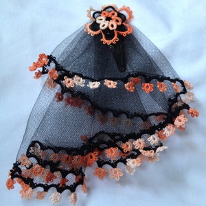 May include: A black tulle veil with orange and cream crocheted trim and a matching flower accent. The veil is attached to a black hair clip.