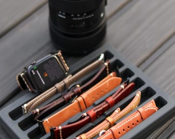 May include: A black Canon camera lens sits above a gray tray holding several leather watch bands in various shades of brown and tan. One watch with a brown band is also visible. The watch bands have white stitching.