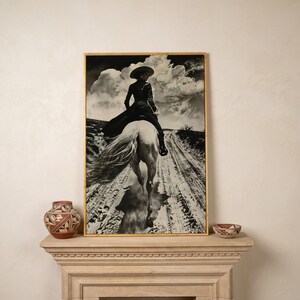 May include: Black and white framed print of a person riding a white horse on a dirt road. The person wears a hat and long coat. The sky is filled with dramatic clouds. Decorative pottery sits on a stone mantel.