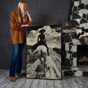 May include: Black and white framed art print depicting a person on horseback riding down a dirt road. The rider is wearing a hat and long coat. The background features a cloudy sky. The art is displayed next to a person wearing a brown shirt and blue jeans.