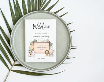 May include: A birthday invitation with the text "Wild One" on a gray plate, surrounded by green leaves. The invitation features illustrations of safari animals and details for Rowan's 1st birthday.