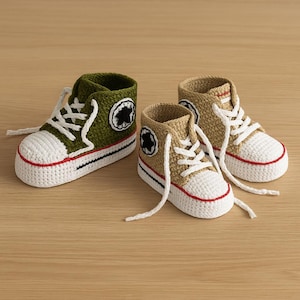 May include: Two pairs of crocheted baby high-top trainers. One pair is olive green, the other is tan. Both have white toes, white laces, and red and black trim. Each shoe has a black and white circular patch on the side.