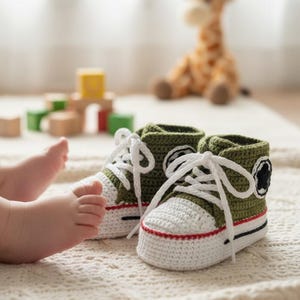 Puede incluir: Un par de zapatillas de bebé de ganchillo hechas a mano en verde oliva, blanco y rojo. Las zapatillas tienen cordones blancos y un detalle de estrella negra en el lateral. Las zapatillas están sobre una superficie texturizada color crema.