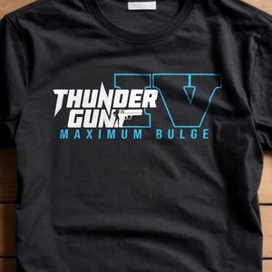 May include: Black t-shirt with the text "THUNDER GUN" in white and a blue graphic. Below the text is "MAXIMUM BULGE" in blue. The shirt has a crew neck and short sleeves.
