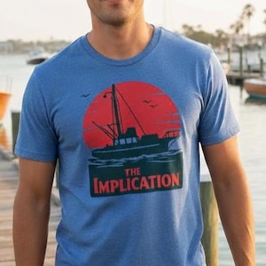 May include: Blue crew neck t-shirt with a graphic of a boat silhouetted against a red sunset, with the text "THE IMPLICATION" in white and red. The shirt is worn by a person standing on a wooden dock.