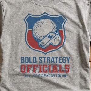 May include: A grey t-shirt with a graphic of a golf ball and whistle inside a shield. The text says "BOLD STRATEGY OFFICIALS" and the tagline "WE'LL SEE IF IT PAYS OFF FOR YOU".