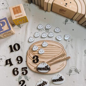May include: Wooden baby milestone set with a dumpling theme. Includes a wooden dumpling steamer, dumplings, numbers, and the words "month" and "year". Also includes wooden blocks and a rainbow stacker.