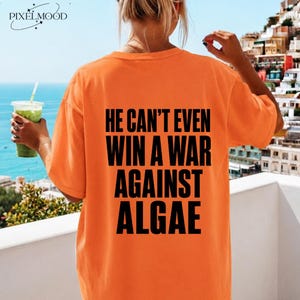 Könnte beinhalten: Ein orangefarbenes Kurzarm-T-Shirt mit fettgedrucktem schwarzem Text auf der Rückseite, der lautet: HE CAN'T EVEN WIN A WAR AGAINST ALGAE. Das Shirt hat einen Rundhalsausschnitt und eine lockere Passform, wobei die Botschaft mittig auf dem Rücken steht.