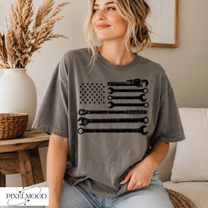 Peut inclure: Un t-shirt gris anthracite à manches courtes avec une impression graphique du drapeau américain composée de silhouettes noires de divers outils à main, dont des clés et un cliquet, disposés en rayures et étoiles.