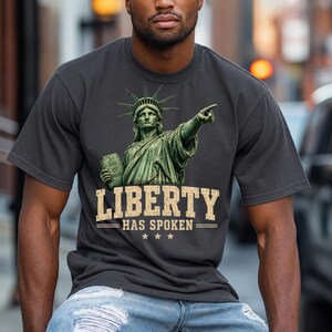 Peut inclure: Un t-shirt gris anthracite à manches courtes avec un motif de la Statue de la Liberté pointant du doigt et tenant une tablette. Sous la statue, le texte LIBERTY HAS SPOKEN est imprimé dans une police beige vieillie avec trois étoiles.