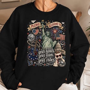 May include: A black sweatshirt featuring a graphic print of the Statue of Liberty, a guitar, a cat wearing heart-shaped sunglasses, and the text PLEDGE ALLEGIANCE TO your hands your team your vibes, along with fireworks and American flag motifs.