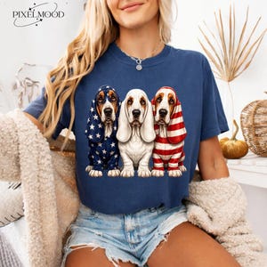 Peut inclure: Un t-shirt bleu marine avec un motif représentant trois chiens Basset Hound. Le chien de gauche porte une capuche bleue à étoiles blanches, celui du centre est blanc, et celui de droite porte une capuche à rayures rouges et blanches.