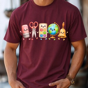 May include: A maroon short-sleeved t-shirt featuring a graphic print of five anthropomorphic school supplies holding hands. The characters include a pink eraser, red scissors, a paint palette, a globe, and a gold hand bell, all with smiling faces.