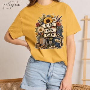 Peut inclure: Un t-shirt à manches courtes jaune moutarde avec une impression graphique représentant un bouquet de tournesols, de marguerites et de lavande. Le motif inclut une bannière avec le texte "STEM COUNT CALM" et des outils de jardinage.