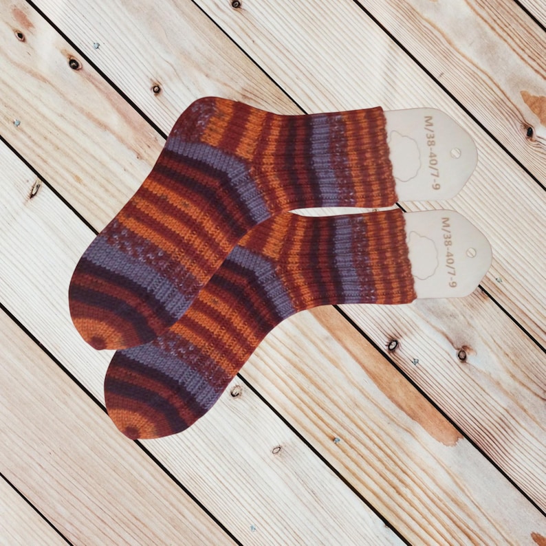 May include: A pair of striped socks in shades of orange, purple, and brown. The socks have a ribbed texture and are displayed on wooden forms. The text "M/38-40/7-9" is printed on the forms. The socks are on a wooden surface.