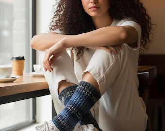 May include: A person wearing white sneakers and blue and gray patterned socks, sitting on a wooden chair. The person is wearing a white outfit and has curly dark hair. A coffee cup sits on a wooden table in the background.