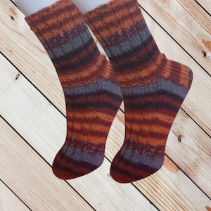 May include: A pair of hand-knitted socks with a striped pattern in shades of orange, burgundy, and gray. The socks are displayed against a wooden background. The socks appear to be made of a soft, warm yarn.