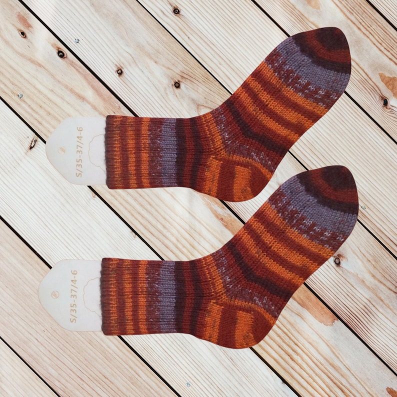 May include: A pair of colorful striped socks with a mix of orange, red, and purple hues. The socks are displayed on a light wooden surface. Each sock has a white tag with text.
