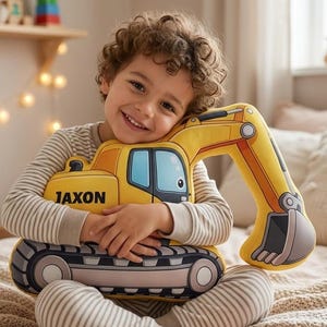 May include: A yellow and black excavator-shaped pillow with the name "JAXON" printed on it. The pillow has a blue window and a detailed design. The excavator is being held by a person wearing striped pajamas.