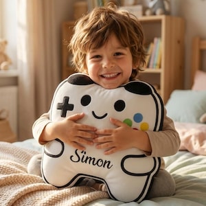 May include: A white and black controller-shaped pillow with the name "Simon" printed on it. The pillow has a smiling face design and colorful buttons. The pillow is being held by a smiling child.