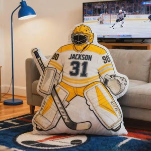 Pode incluir: Uma almofada em forma de guarda-redes de h&oacute;quei em branco e amarelo, com o nome "Jackson" e o n&uacute;mero "31" na camisola. A almofada tem um stick de h&oacute;quei preto e est&aacute; sobre um tapete estampado.