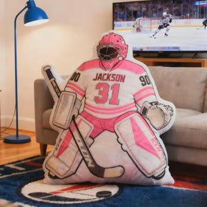 Pode incluir: Uma almofada de guarda-redes de h&oacute;quei branca e rosa, com o nome "Jackson" e o n&uacute;mero "31". A almofada tem a forma de um guarda-redes com equipamento completo, com capacete, prote&ccedil;&otilde;es e um stick de h&oacute;quei. A almofada est&aacute; sobre um tapete estampado.