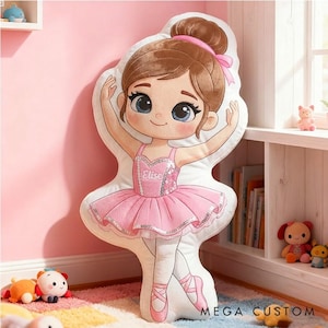 Op de afbeelding: Een groot, pluche kussen in de vorm van een cartoon ballerina. Het kussen heeft een roze tutu, een lijfje met pailletten en balletschoenen. De ballerina heeft bruin haar, blauwe ogen en de naam "Elise" op het lijfje. Het kussen ligt op een kleurrijk tapijt.