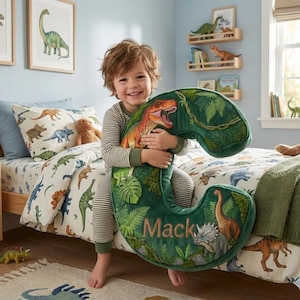 Op de afbeelding: Een kind houdt een groot, groen dinosaurusvormig kussen vast met de naam "Mack" erop geborduurd. Het kussen heeft illustraties van verschillende dinosaurussen en tropisch gebladerte. Het beddengoed en de kunst aan de muur in de kamer hebben ook dinosaurusthema's.