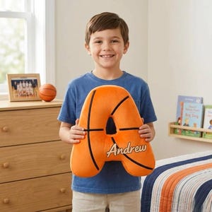 Op de afbeelding: Een oranje basketbal-thema letter "A" kussen met zwarte strepen en de naam "Andrew" in script. Een kind in een blauw shirt houdt het kussen vast. Een basketbal en een ingelijste foto staan op de achtergrond.