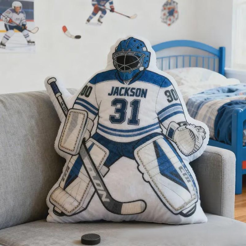 Pode incluir: Uma almofada em forma de guarda-redes de h&oacute;quei, branca e azul, com o nome "Jackson" e o n&uacute;mero "31" na camisola. A almofada est&aacute; num sof&aacute; cinzento, com um disco de h&oacute;quei &agrave; frente. Decora&ccedil;&atilde;o de parede com tema de h&oacute;quei.