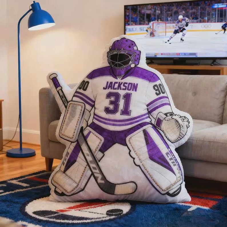 Pode incluir: Uma almofada de pel&uacute;cia em forma de goleiro de h&oacute;quei com uniforme branco e roxo. A almofada apresenta o nome "Jackson" e o n&uacute;mero "31". O goleiro est&aacute; usando capacete e protetores, com um taco e uma luva de h&oacute;quei.