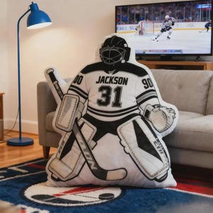 Pode incluir: Uma almofada de guarda-redes de h&oacute;quei em branco, preto e cinza, com o nome "JACKSON" e o n&uacute;mero "31". A almofada tem a forma de um guarda-redes com equipamento completo, incluindo capacete, prote&ccedil;&otilde;es e um stick de h&oacute;quei.