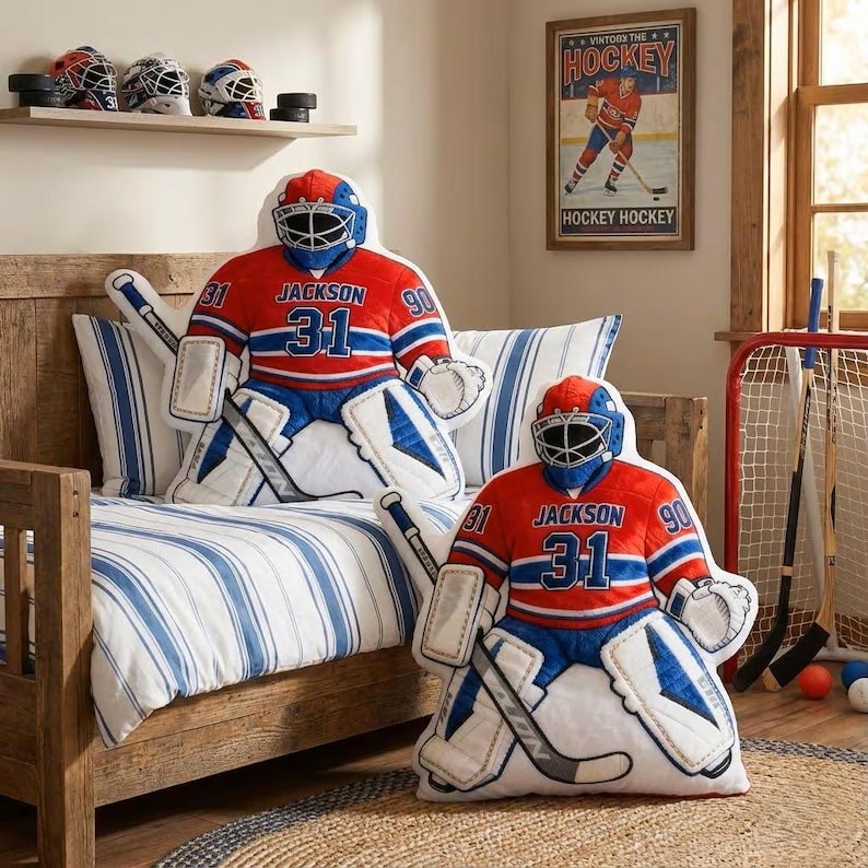 Pode incluir: Duas almofadas em forma de guarda-redes de h&oacute;quei em vermelho, azul e branco, com o nome "Jackson" e o n&uacute;mero "31" na frente. As almofadas est&atilde;o numa cama com roupa de cama &agrave;s riscas azuis e brancas. Um cartaz de h&oacute;quei vintage est&aacute; pendurado na parede.