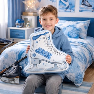 May include: A young person holds a decorative pillow shaped like a white ice skate with blue accents and the name "JACK" in blue. The pillow features snowflake designs. The background includes a blue bedspread and other winter-themed decor.
