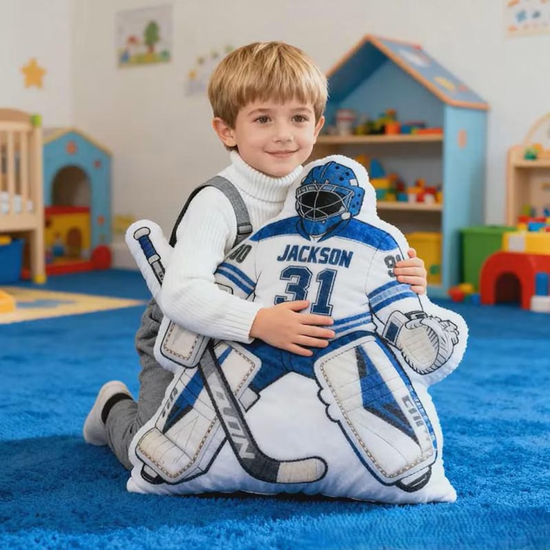 Pode incluir: Uma almofada de guarda-redes de h&oacute;quei em branco, azul e cinza. A almofada apresenta o nome "Jackson" e o n&uacute;mero "31" na camisola. A almofada tem a forma de um jogador de h&oacute;quei e inclui um stick e prote&ccedil;&otilde;es.