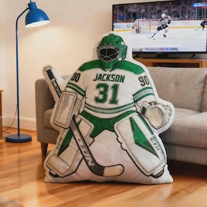 Pode incluir: Uma almofada em forma de guarda-redes de h&oacute;quei, em branco e verde, com o nome "Jackson" e o n&uacute;mero "31". A almofada tem um capacete e prote&ccedil;&otilde;es verdes e est&aacute; colocada num sof&aacute; claro.